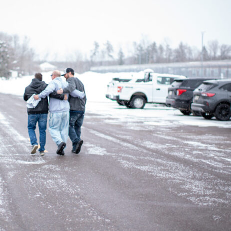 A man wearing a grey sweatshirt and jeans is surrounded by his brother and adult son while walking through a snowy parking lot.