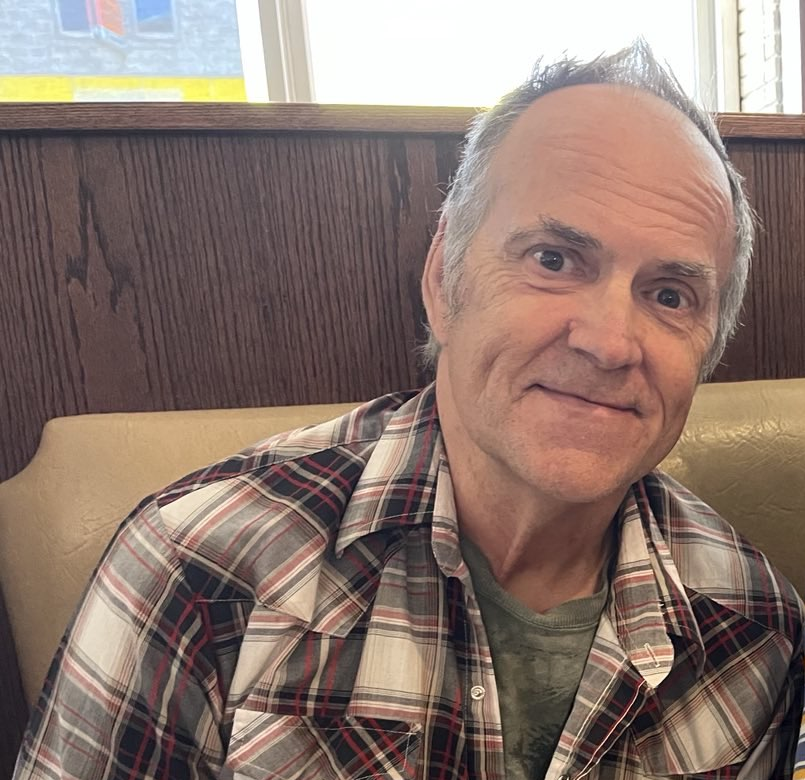 An older man with grey hair sits in a cafe  booth wearing a plaid shirt
