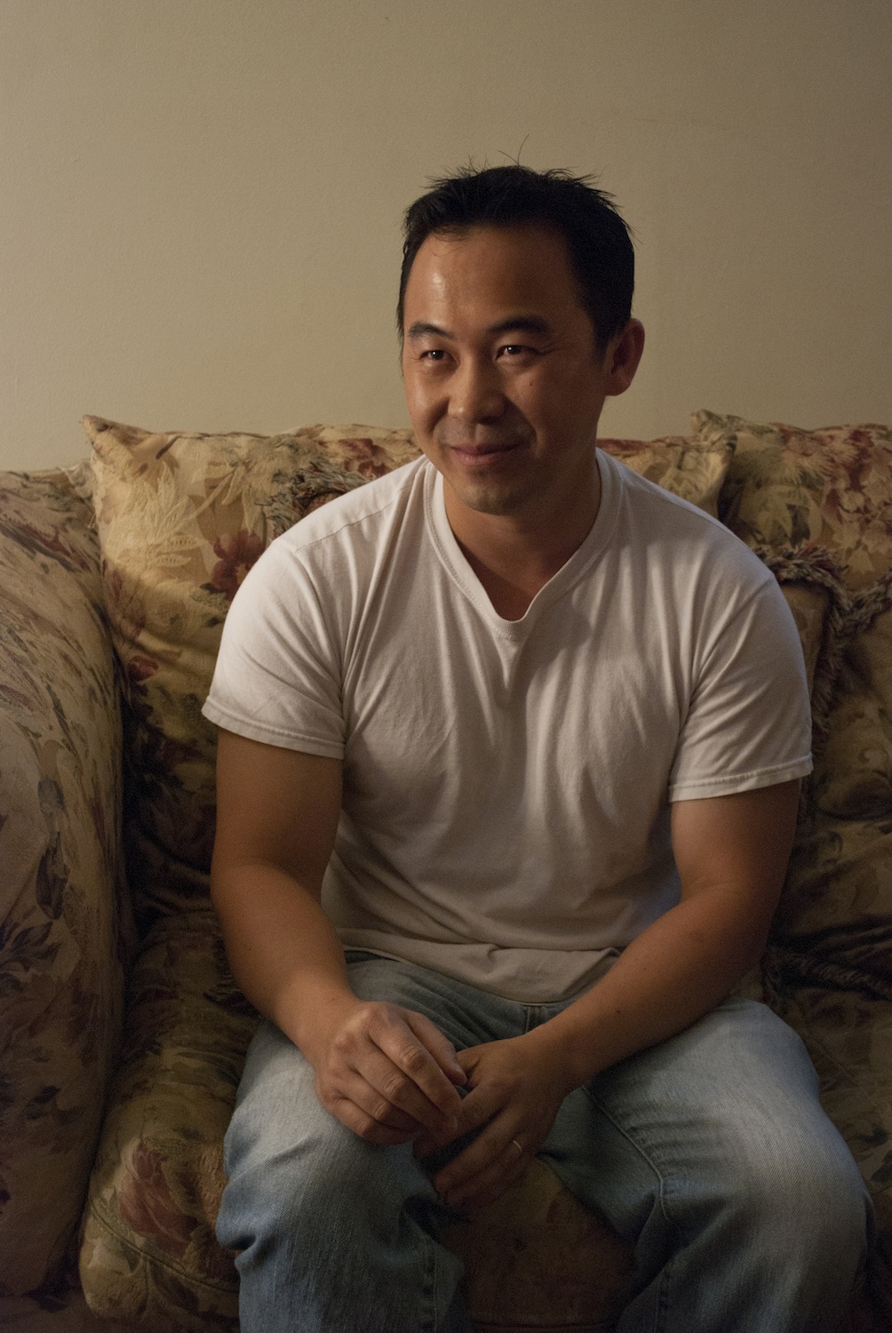 An Asian man sits on a floral couch wearing a white tee-shirt.