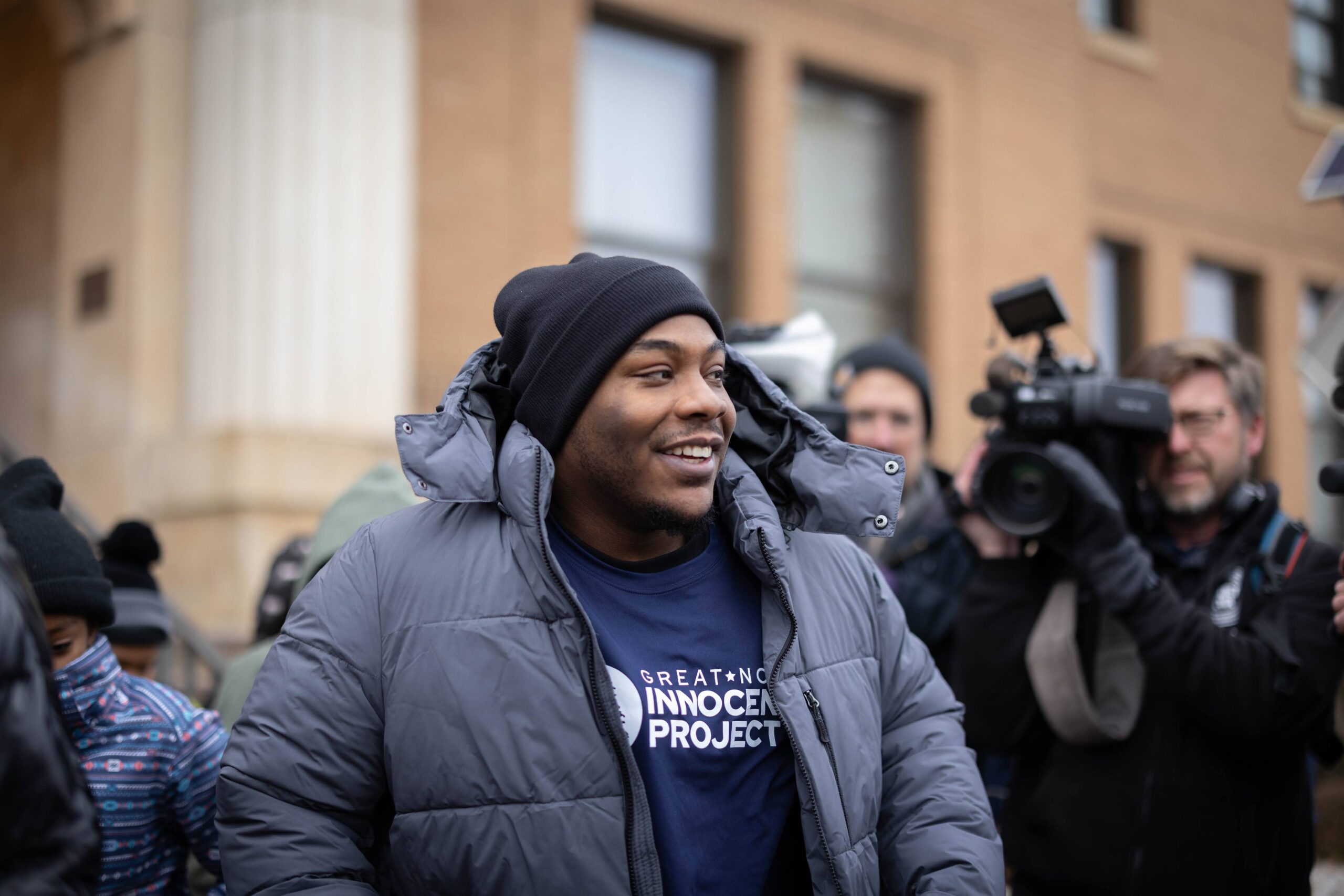 A young Black man wearing a black beanie, winter coat, and Great North Innocence project hoody looks on at a crowd of supporters and media