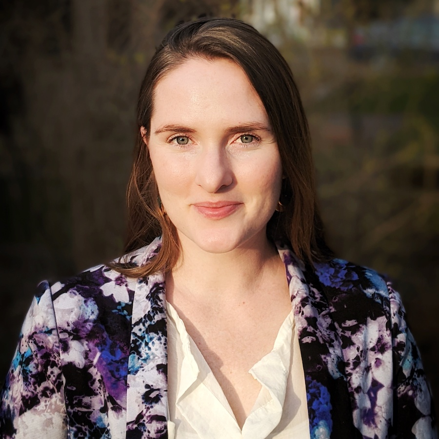 A woman with light brown hair wears a purple floral blazer and smiles outside