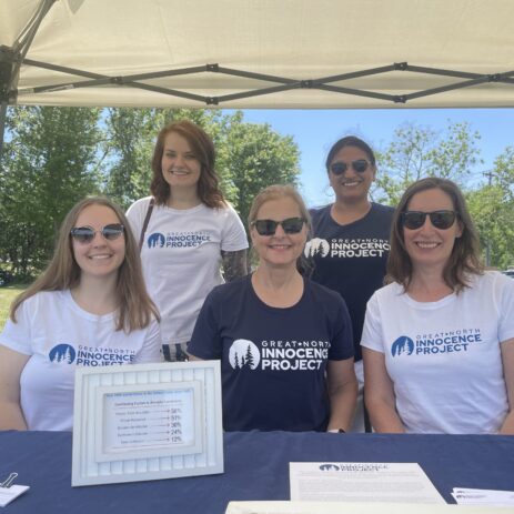 A group of five women wearing Great North Innocence Project shirts pose in front of a table advertising the organization's services.