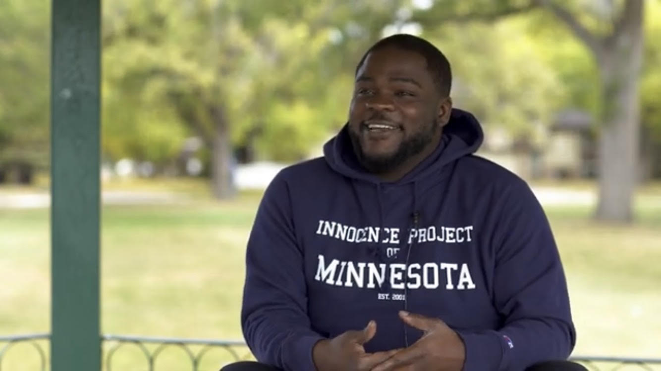A young Black man wearing an Innocence Project of Minnesota sweatshirt speaks at a park