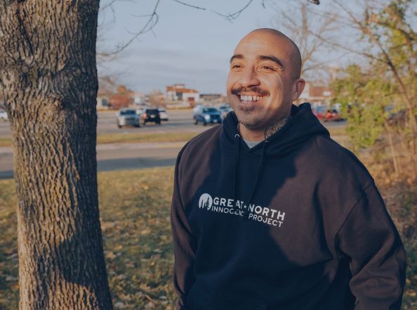 A middle age, Hispanic man wearing a navy blue Great North Innocence Project sweatshirt smiles in front of tree.