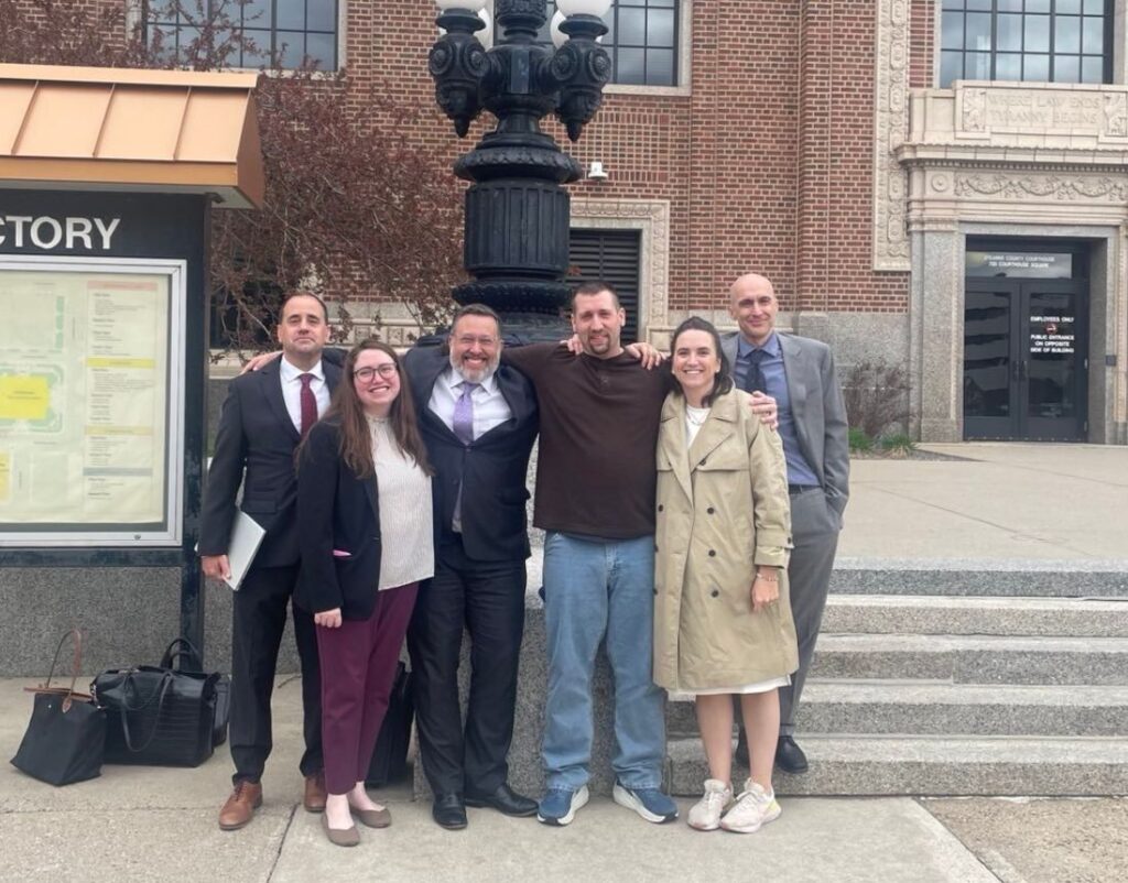 A man wearing a brown shirt and jeans is surrounded by his attorneys standing in front of a courthouse