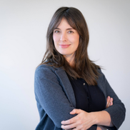 A woman with light brown hair wears a grey blazer and has crossed arms
