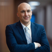 A bald man wears a navy suit jacket and blue striped tie
