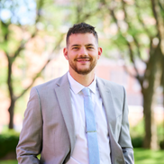 A young man in a light grey suit jacket and blue tie smiles outside