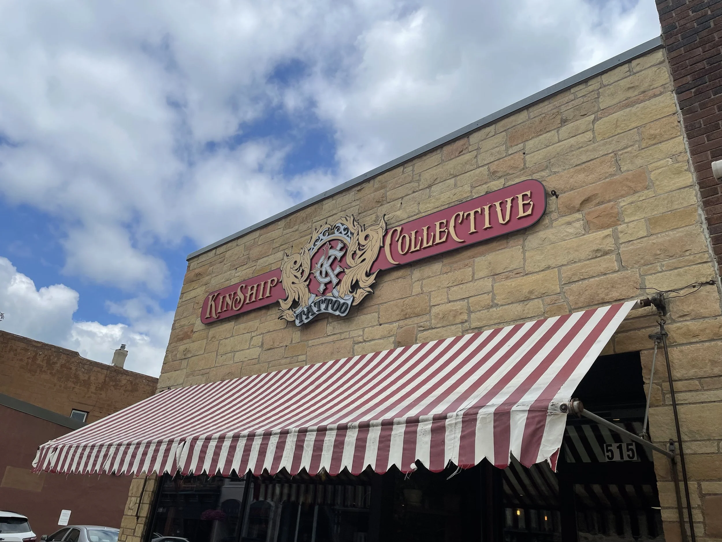 A building with a red and gold sign reading "Kinship Collective Tattoo" above a white and red striped banner