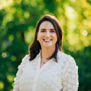 A white woman in her 30's with brown hair and a white blouse smiles in front of sun drenched trees